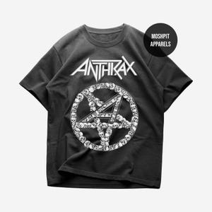 May include: Black t-shirt with the white band name "ANTHRAX" above a white pentagram design surrounded by skulls. The shirt has a crew neck and short sleeves. The brand name "MOSHPIT APPARELS" is printed on a black circle.