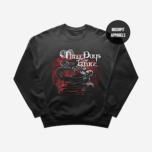 May include: Dark gray sweatshirt featuring a graphic of a snake coiled around a skull, with the band name "Three Days Grace" in white lettering on a red background. The sweatshirt has a crew neck and long sleeves. The brand name "Moshpit Apparels" is in a black circle.