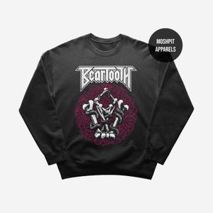 May include: Black sweatshirt with the band name "Beartooth" in white, above a graphic of a skeleton and snakes within a pink rose design. The sweatshirt has a crew neck and long sleeves. The brand "Moshpit Apparels" is printed on a black circle.