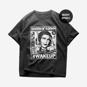 Könnte beinhalten: Dunkelgraues T-Shirt mit einem Schwarz-Weiß-Grafikdesign. Das Design enthält den Text "SYSTEM OF A DOWN", "RESIST suffering #WAKEUP" und das Bild einer Frau mit einer Pistole.