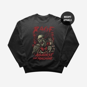 May include: Dark gray sweatshirt featuring a red graphic of a skeleton holding a microphone, with the words "RAGE AGAINST THE MACHINE" in red. The sweatshirt has a crew neck and long sleeves. The brand name "MOSHPIT APPARELS" is in a black circle.