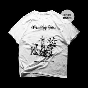 May include: White t-shirt featuring a black graphic of a band logo and a scene of figures with birds. The text "Three Days Grace" is above the graphic, and "LIFE STARTS NOW" is below. The shirt has a crew neck and short sleeves.