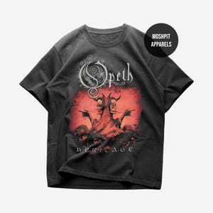 Puede incluir: Camiseta gris oscuro con un diseño gráfico de la banda "Opeth" en escritura blanca sobre un fondo rojo con dos figuras con cuernos. La palabra "heritage" está impresa debajo.