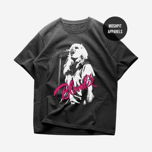 May include: Dark gray t-shirt featuring a black and white image of a singer with the word "Blondie" in pink script. The shirt has a crew neck and short sleeves. The brand name "Moshpit Apparels" is printed on a black circle.