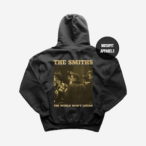 Op de afbeelding: Donkergrijze hoodie met een sepia-getinte afbeelding van een groep mensen. De tekst "THE SMITHS" staat boven de afbeelding en "THE WORLD WON'T LISTEN" eronder. Een zwarte cirkel met "MOSHPIT APPARELS" staat rechts.