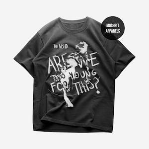 May include: Dark grey t-shirt featuring the white text "ARE WE TOO YOUNG FOR THIS?" and "THE NBHD". The shirt has a crew neck and short sleeves. A black circle with "MOSHPIT APPARELS" is on the upper right.