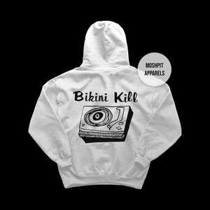 Może przedstawiać: Jasnoszara bluza z kapturem z czarną grafiką gramofonu i napisem "Bikini Kill" z tyłu. Bluza z kapturem ma również napis "Moshpit Apparels" w prawym górnym rogu.