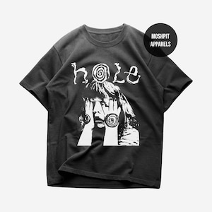 Puede incluir: Camiseta negra con un gráfico blanco. El diseño presenta la palabra "hole" sobre una ilustración de una persona cubriéndose los ojos con las manos. El gráfico incluye elementos en espiral. La camiseta tiene cuello redondo y mangas cortas.