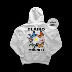 May include: A white hoodie featuring the word "CLAIRO" above a butterfly design with blue, orange, and pink butterflies. Below the graphic is the word "IMMUNITY" and a list of song titles. The hoodie has a hood and long sleeves.