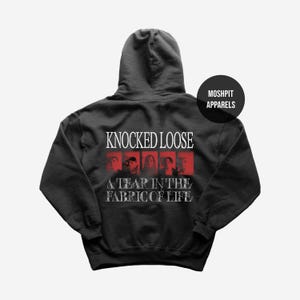 Puede incluir: Sudadera con capucha gris oscuro con el texto "KNOCKED LOOSE" en blanco, encima de una fila de cuadrados rojos con imágenes, y el texto "A TEAR IN THE FABRIC OF LIFE". Un círculo negro con "MOSHPIT APPARELS" está en la parte superior derecha.