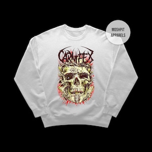 May include: White sweatshirt with a detailed skull graphic and the word "CARNIFEX" in red. The skull design has green, yellow, and red accents. The sweatshirt has a crew neck and long sleeves. The brand name "MOSHPIT APPARELS" is printed on a white circle.