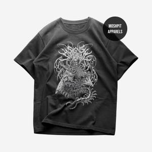 Puede incluir: Camiseta gris oscuro con un gráfico blanco detallado de una figura esquelética con alas, rodeada de diseños abstractos intrincados. La camiseta incluye el texto "MOSHPIT APPARELS" en un logotipo circular. El diseño tiene un tema gótico o metal.