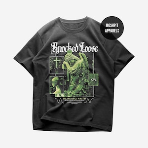 Puede incluir: Camiseta gris oscuro con un gráfico verde de una estatua de ángel llorando, una cruz y el texto "Knocked Loose" en fuente gótica. La camiseta también incluye el texto "Blinding Faith" y "K/L".
