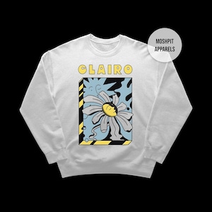 May include: White sweatshirt with the word "Clairo" in yellow block letters above a daisy graphic. The daisy has a face and is set against a blue and black background within a yellow and black striped frame. The brand name "Moshpit Apparels" is in a white circle.
