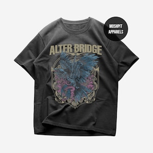 Op de afbeelding: Donkergrijs T-shirt met de bandnaam "ALTER BRIDGE" boven een gedetailleerde afbeelding van een raaf met blauwe en paarse accenten. Het ontwerp bevat een slang en bloemelementen. Het merklogo "MOSHPIT APPARELS" staat in een zwarte cirkel.