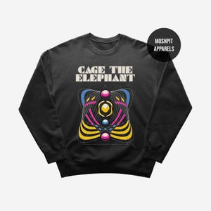 Può includere: Felpa girocollo nera con la scritta "CAGE THE ELEPHANT" sopra un design geometrico colorato. Il design include linee e sfere gialle, blu, rosa e viola. Il marchio "MOSHPIT APPARELS" è su un cerchio nero.
