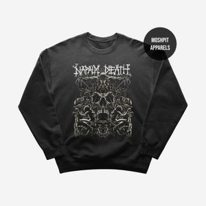 Peut inclure: Sweat-shirt gris foncé avec un motif de crâne et de figures, et le nom du groupe "NAPALM DEATH" en blanc. Le sweat-shirt porte également le logo "MOSHPIT APPARELS".
