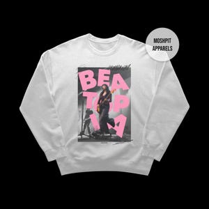 May include: White crew neck sweatshirt with a pink graphic that reads "BEAT TOP IN A" and a photo of a person playing a guitar. The sweatshirt has long sleeves and a round neckline. The brand name "MOSHPIT APPARELS" is printed on a white circle.