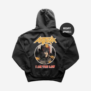 May include: Dark grey hooded sweatshirt with the band name "Anthrax" in gold lettering. Below is a circular graphic of a figure in a helmet, with the text "I AM THE LAW" in red. The sweatshirt also has a "Moshpit Apparels" logo.