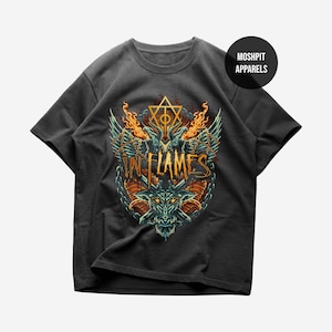 May include: Dark gray t-shirt featuring a detailed graphic design. The design includes a dragon-like creature, flames, wings, and the text "IN FLAMES" in an ornate font. A circular logo with the text "MOSHPIT APPARELS" is in the upper right corner.
