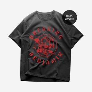 May include: Dark gray t-shirt with a red graphic featuring a skull, lightning bolt, and the words "BREAKING BENJAMIN" in a circular design. The shirt has a crew neck and short sleeves. The brand "MOSHPIT APPARELS" is printed on a black circle.