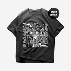Puede incluir: Una camiseta gris oscuro con un diseño gráfico en blanco y negro. El diseño incluye el texto "THE NBHD" y un logotipo, junto con ilustraciones de manos, plumas y mariposas. La camiseta tiene mangas cortas y cuello redondo.