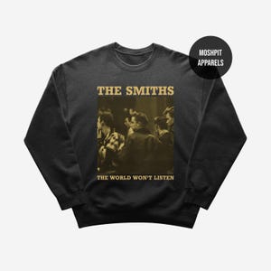 Op de afbeelding: Donkergrijze sweatshirt met de bandnaam "THE SMITHS" boven een sepia-kleurige afbeelding van mensen. Onder de afbeelding staat de tekst "THE WORLD WON'T LISTEN". Een zwarte cirkel met "MOSHPIT APPARELS" staat in de rechterbovenhoek.