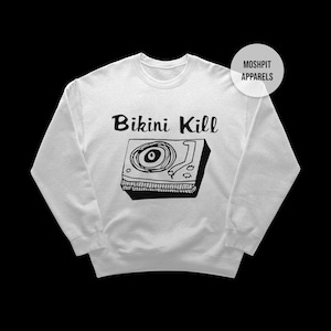 Może przedstawiać: Biała bluza z napisem "Bikini Kill" nad czarno-białą ilustracją gramofonu. Okrągłe logo z napisem "Moshpit Apparels" znajduje się w prawym górnym rogu.
