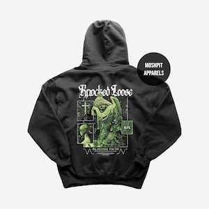 Puede incluir: Sudadera con capucha negra con un diseño gráfico en la espalda. El diseño presenta una estatua de ángel, una cruz y las palabras "Knocked Loose" y "Blinding Faith" en blanco. También es visible la marca "Moshpit Apparels".