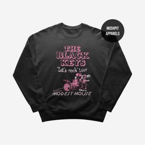 Può includere: Felpa girocollo nera con grafica rosa. La grafica presenta il testo "THE BLACK KEYS 'Let's rock' tour MODEST MOUSE" con un'illustrazione della band. Un cerchio nero con la scritta "MOSHPIT APPARELS" si trova in alto a destra.