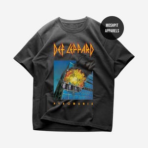 Könnte beinhalten: Dunkelgraues Def Leppard Pyromania T-Shirt. Das Shirt zeigt das Bandlogo in Gelb und Orange über einer quadratischen Grafik einer Uhr und eines Gebäudes. Das Wort "PYROMANIA" ist darunter in Gelb gedruckt.