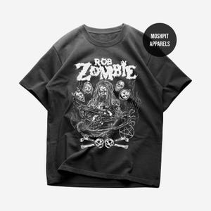 May include: Black t-shirt featuring a graphic of Rob Zombie with skulls, bones, and a pentagram. The text "ROB ZOMBIE" is displayed above the graphic. The shirt has short sleeves and a crew neck. The brand name "MOSHPIT APPARELS" is printed on a black circle.