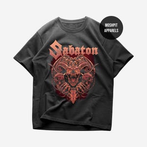 Könnte beinhalten: Dunkelgraues T-Shirt mit einem detaillierten rot-orangen Motiv eines gehörnten Schädels und dem Wort "Sabaton" in Rot. Das Shirt hat einen Rundhalsausschnitt und kurze Ärmel. Die Marke "Moshpit Apparels" ist auf einem schwarzen Kreis aufgedruckt.