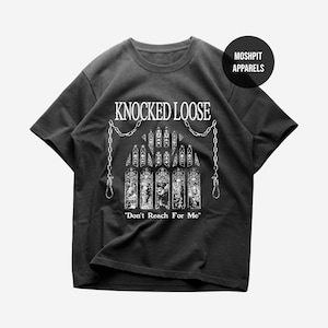 Puede incluir: Camiseta negra con el texto blanco "KNOCKED LOOSE" sobre un gráfico detallado de vidrieras y cadenas. La frase "Don't Reach For Me" está impresa debajo. Un logotipo redondo con "MOSHPIT APPARELS" está en la parte superior derecha.