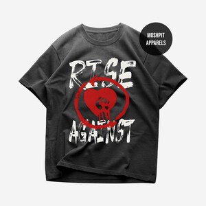 May include: Dark gray t-shirt with the words "RISE AGAINST" in white, distressed lettering. A red heart with a skull graphic is centered on the front. The shirt has short sleeves and a crew neck.