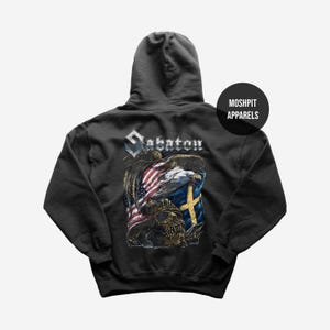 Könnte beinhalten: Dunkelgrauer Kapuzenpullover mit einem grafischen Design mit dem Wort "Sabaton", einem Adler, der amerikanischen Flagge und der schwedischen Flagge. Am unteren Rand des Designs befindet sich ein Totenkopf. Der Kapuzenpullover hat eine Kapuze und ein rundes Etikett mit der Aufschrift "Moshpit Apparels".
