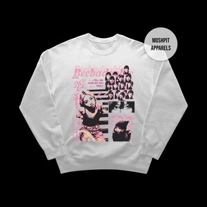 May include: White sweatshirt featuring a pink and black graphic design. The design incorporates the text "Beebadobee" and "Tell Me How Do You Feel?" alongside images of a woman. The sweatshirt has a crew neck and long sleeves.