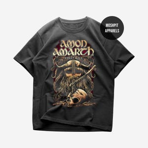 May include: Dark gray t-shirt featuring a Viking warrior skull design with the text "AMON AMARTH FIRST KILL" in gold and red. The design includes a horned helmet, beard, and a skull. The shirt is from Moshpit Apparels.