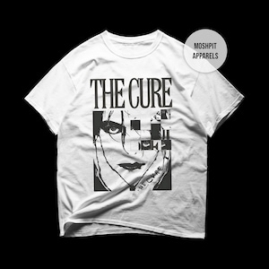 May include: White t-shirt featuring a black and white graphic of a face with the text "THE CURE" above and "THE CURE" below the face. The shirt has short sleeves and a crew neck. The brand name "MOSHPIT APPARELS" is on the upper right.