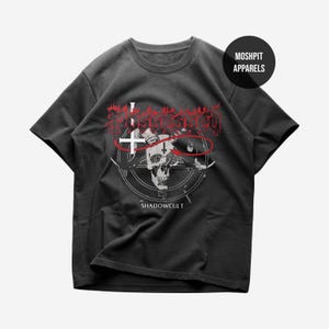 May include: Dark gray t-shirt with a graphic design featuring a skull, a cross, and the text "SHADOWCULT". The design is in red and white. The shirt also has the text "MOSHPIT APPARELS" in a black circle.