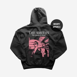 Op de afbeelding: Een zwarte hoodie met een roze afbeelding van twee mannen en de tekst "THE SMITHS The Queen Is Dead". De hoodie heeft ook de tekst "MOSHPIT APPARELS" in een zwarte cirkel.