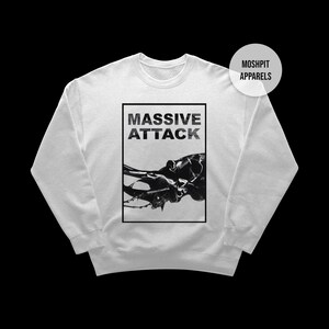 Könnte beinhalten: Ein weißes Sweatshirt mit dem Text "MASSIVE ATTACK" in einem schwarzen Rechteck und einer schwarz-weißen Grafik. Das Sweatshirt hat einen Rundhalsausschnitt und lange Ärmel. Ein weißer Kreis mit "MOSHPIT APPARELS" befindet sich in der oberen rechten Ecke.