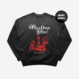 May include: Black crewneck sweatshirt featuring the band name "Three Days Grace" in white script above a red graphic. The text "LIFE STARTS NOW" is below the graphic. A black circle with "MOSHPIT APPARELS" is in the upper right corner.