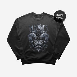 May include: Dark gray sweatshirt featuring a detailed black and white graphic of a snarling wolf with large horns, and the text "IN FLAMES" above. A black circle with the text "MOSHPIT APPARELS" is in the upper right corner. The sweatshirt is a crew neck style.