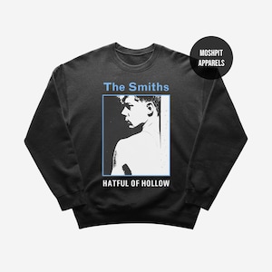 Op de afbeelding: Donkergrijze sweatshirt met een zwart-wit afbeelding van een persoon, de tekst "The Smiths" en "Hatful of Hollow" in lichtblauw. Een zwarte cirkel met "Moshpit Apparels" staat in de rechterbovenhoek.