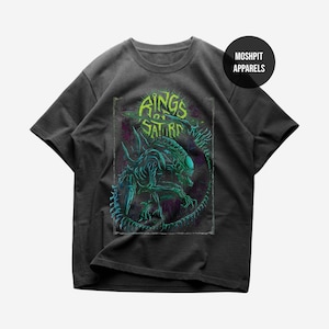 Op de afbeelding: Donkergrijs t-shirt met een neon groen en blauw alien-ontwerp en de tekst "Rings of Saturn". Het ontwerp staat in een ovale, verweerde lijst. Het shirt heeft korte mouwen en een ronde hals.