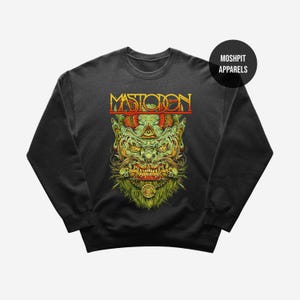 May include: Dark gray sweatshirt featuring a detailed graphic of a green monster with a beard, fangs, and a third eye. The word "MASTODON" is printed in red and yellow above the monster. A black circle with "MOSHPIT APPARELS" is in the upper right.