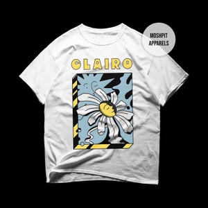 May include: White t-shirt with a daisy graphic and the word "Clairo" in yellow block letters. The daisy has white petals and a yellow centre, set against a light blue background with black accents. The shirt has a crew neck.