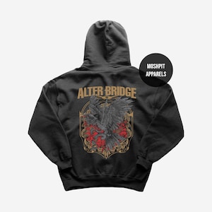 Op de afbeelding: Donkergrijze hoodie met een grote afbeelding op de achterkant. De afbeelding toont een zwarte vogel met uitgespreide vleugels, een rood bloemenpatroon en de tekst "ALTER BRIDGE" in gouden letters. De merknaam "MOSHPIT APPARELS" is ook zichtbaar.