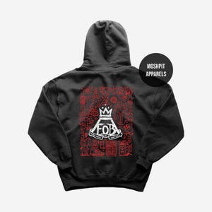 Peut inclure: Sweat à capuche gris foncé avec un motif graphique rouge et blanc. Le motif présente les lettres "FOB" avec une couronne, entourées de diverses illustrations et du texte "SAVE ROCK AND ROLL". Le sweat à capuche a une capuche et la marque "MOSHPIT APPARELS" est imprimée.
